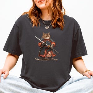 May include: Dark gray t-shirt featuring a cartoon cat dressed as a samurai, holding a katana. The cat is wearing red and black armor. The design is centered on the shirt, with a detailed illustration of the cat and its attire.
