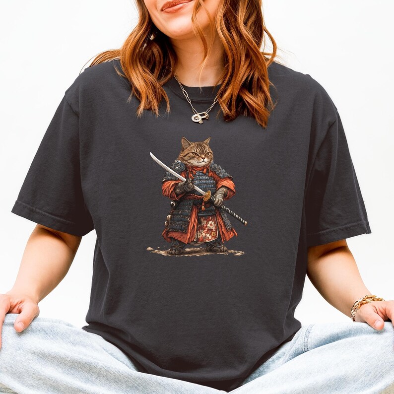 May include: Dark gray t-shirt featuring a cartoon cat dressed as a samurai, holding a katana. The cat is wearing red and black armor. The design is centered on the shirt, with a detailed illustration of the cat and its attire.