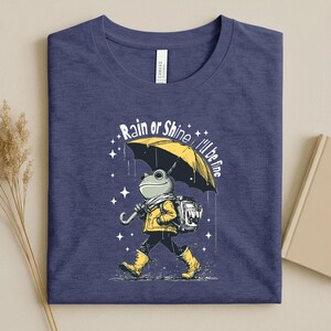 May include: Navy blue t-shirt featuring a whimsical illustration of a frog walking in the rain under a yellow umbrella. The frog wears a yellow jacket, boots, and a backpack. Text on the shirt reads "Rain or Shine, I'll be fine."