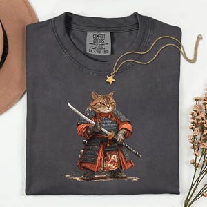 May include: Dark grey t-shirt featuring a cartoon cat dressed as a samurai, holding a sword. The cat is wearing red and black armor. A gold star pendant necklace and a Comfort Colors tag are also visible.