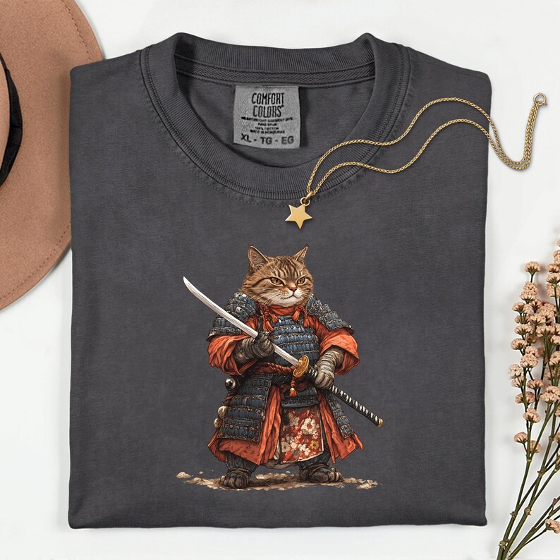 May include: Dark grey t-shirt featuring a cartoon cat dressed as a samurai, holding a sword. The cat is wearing red and black armor. A gold star pendant necklace and a Comfort Colors tag are also visible.