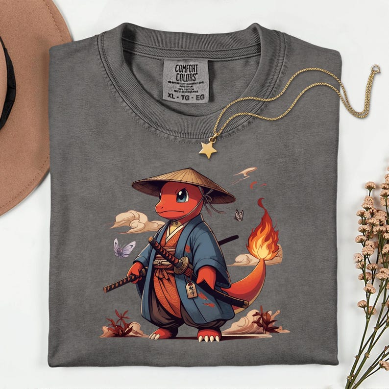 May include: A gray Comfort Colors t-shirt featuring a cartoon character in a samurai outfit, holding two swords. The character is red with a flame tail, wearing a straw hat and blue kimono. A gold star necklace is draped on the shirt.