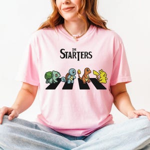 May include: Pale pink t-shirt with the words "The Starters" above a graphic of four cartoon characters crossing a black and white crosswalk. The characters are Bulbasaur, Squirtle, Charmander, and Pikachu.