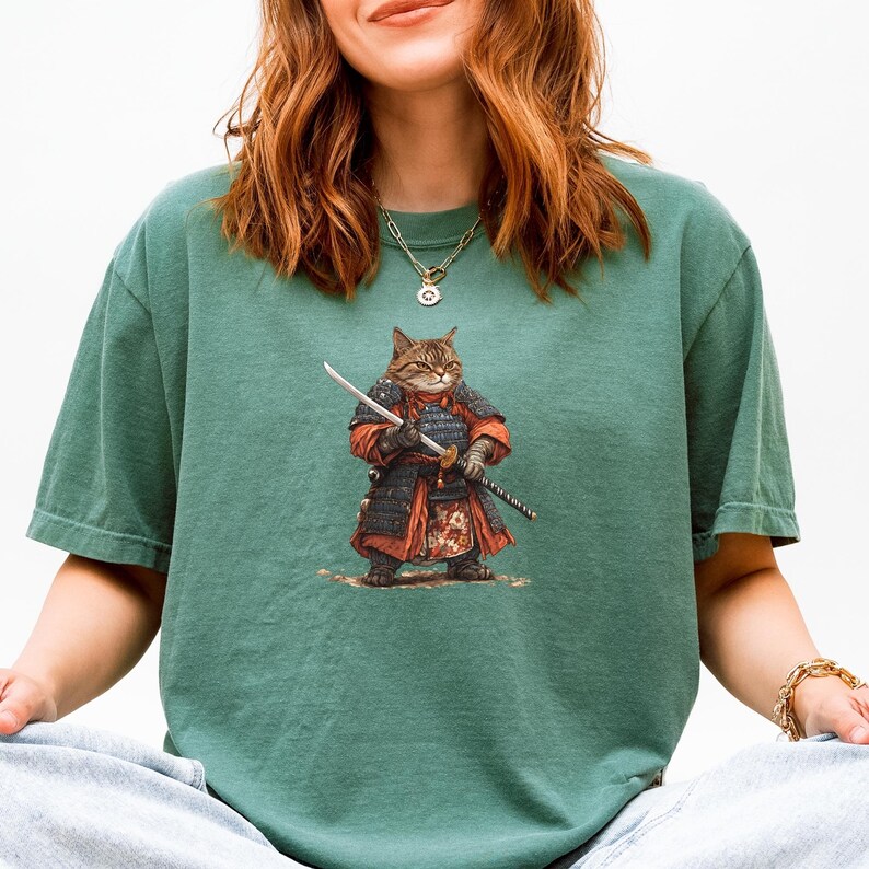 May include: Green t-shirt featuring a detailed illustration of a cat dressed as a samurai, holding two swords. The cat is wearing traditional samurai armor in shades of red, black, and brown. The shirt is a solid green color.