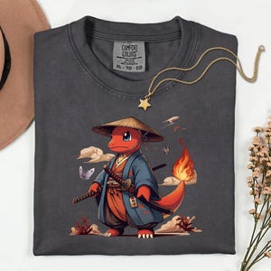 May include: Dark grey t-shirt featuring a cartoon character in a samurai outfit. The character is red with a flame tail, wearing a blue kimono and straw hat, holding two swords. The shirt has a Comfort Colors tag and a gold star necklace.