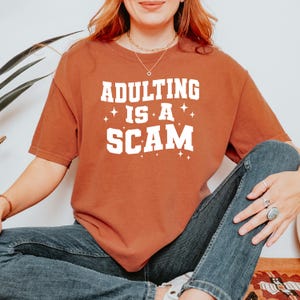 May include: Rust-colored short-sleeved t-shirt with the phrase "ADULTING IS A SCAM" in white block letters. The text is accented with small star shapes. The shirt is worn with blue jeans.