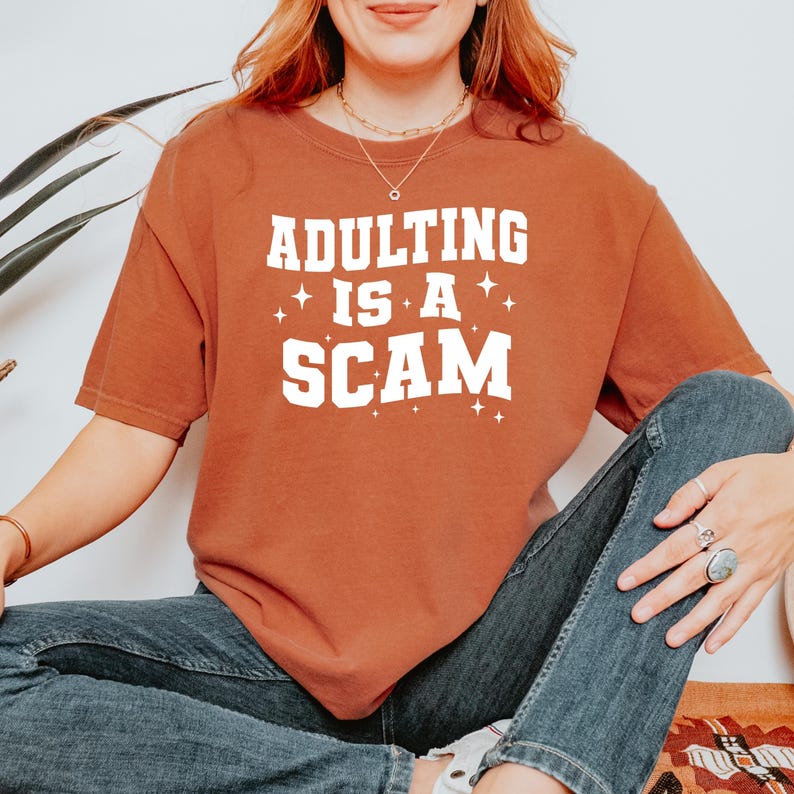 May include: Rust-colored short-sleeved t-shirt with the phrase "ADULTING IS A SCAM" in white block letters. The text is accented with small star shapes. The shirt is worn with blue jeans.