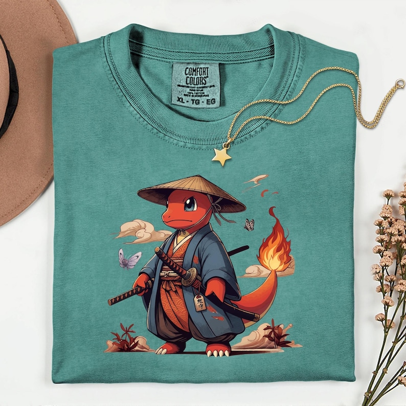 May include: Teal Comfort Colors t-shirt featuring a cartoon character in a samurai outfit, complete with a straw hat and swords. The character is red with a flame tail. A gold star pendant necklace rests on the shirt. The shirt has a tag that says XL-TG-EG.
