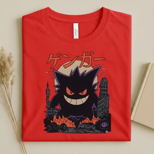 May include: Red t-shirt featuring a black cartoon monster with orange eyes and a wide grin, set against a cityscape with flames. Japanese text is above the monster. The shirt is made of a soft material.