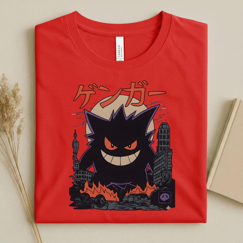 May include: Red t-shirt featuring a black cartoon monster with orange eyes and a wide grin, set against a cityscape with flames. Japanese text is above the monster. The shirt is made of a soft material.
