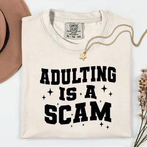 May include: Cream-colored t-shirt with the phrase "ADULTING IS A SCAM" in bold black letters. The shirt features a Comfort Colors tag and is accessorized with a gold necklace with a star pendant. A brown hat and flowers are in the background.