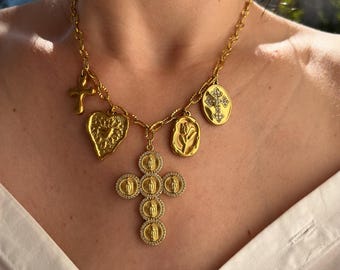 Op de afbeelding: Een goudkleurige ketting met meerdere religieuze bedels. De bedels omvatten een kruis, een hart, een roos en een kruisvormige hanger met ingebedde figuren. De ketting wordt gedragen over een wit overhemd.