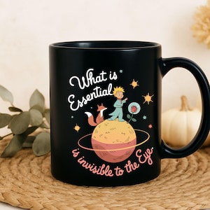 May include: Black ceramic mug featuring a whimsical illustration from "The Little Prince." The design includes the text "What is Essential is invisible to the Eye." The artwork depicts the Little Prince, a fox, a rose, and a planet with stars.