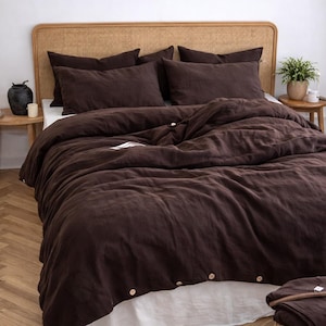 May include: A bed dressed with a dark brown linen duvet cover and pillowcases. The bed features a woven headboard and a white bed skirt. A small side table holds a plant and a decorative vase.