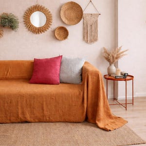 Op de afbeelding: Een gezellige woonkamer met een oranje bank met rode en grijze kussens. Decoratieve wanddecoraties zijn onder meer een spiegel, geweven borden en een macramé stuk. Een bijzettafel met een vaas met droogbloemen en een hanglamp van riet.