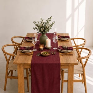 Op de afbeelding: Een rechthoekige houten eettafel met vier licht houten stoelen met gevlochten zittingen. Een donkerrode linnen tafelloper ligt in het midden van de tafel, versierd met een groene vaas met eucalyptus, een witte stompkaars en servies.