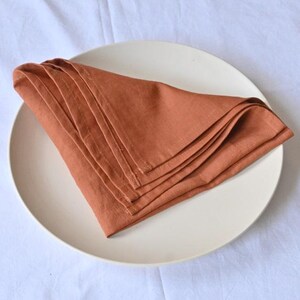 May include: A stack of folded, rust-colored linen napkins rests on a cream-colored plate. The plate is centered on a white, textured tablecloth. The napkins are neatly arranged, showcasing their soft texture and warm hue.