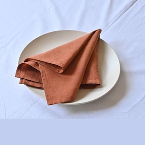 May include: A folded rust-colored linen napkin rests on a cream-colored plate. The plate is centered on a white, slightly wrinkled tablecloth. The napkin is folded into a triangle shape, with multiple layers visible.
