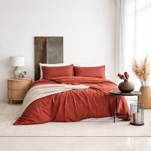 以下が含まれることがあります： 赤錆色の寝具が特徴の寝室のシーン。掛け布団と枕2つが含まれます。ベージュのブランケットがベッドに掛けられています。木製のヘッドボードと、ランプ付きのベッドサイドテーブルが見えます。