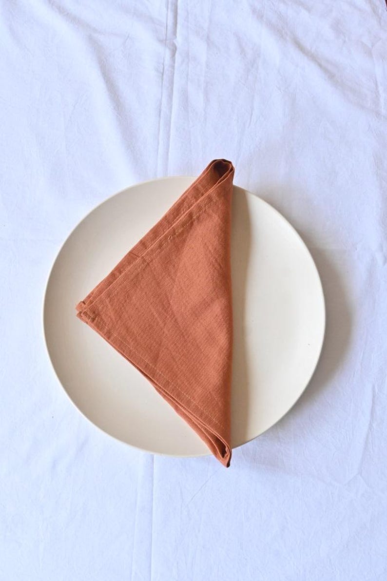 May include: A folded, terracotta-coloured linen napkin sits on a cream-coloured plate. The napkin is folded into a triangle shape, with visible texture. The plate rests on a white, slightly wrinkled tablecloth, creating a simple, elegant setting.
