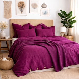 May include: A bedroom scene featuring a bed with a deep magenta duvet cover and matching pillows. A woven headboard and a woven rug add texture. Decorative wall art and a large plant complete the cozy, inviting space.