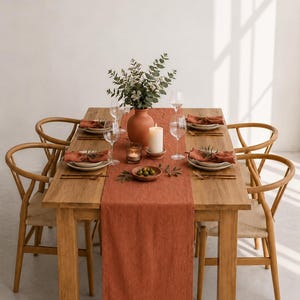 May include: A rectangular wooden dining table set with four light wood chairs, a terracotta-colored linen table runner, white plates, rust-colored napkins, clear wine glasses, a terracotta vase with greenery, a white pillar candle, and olives.