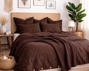 May include: A dark brown bedding set featuring a duvet cover with visible light-coloured buttons along the bottom edge, matching pillowcases, and a textured waffle-weave throw blanket draped over the bed, creating a cohesive and layered appearance.