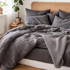 May include: A bed with gray linen bedding, including a duvet cover, pillows, and a waffle-knit throw blanket. The headboard is made of woven rattan. A potted plant sits on a small wooden side table near a window.