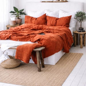 以下が含まれることがあります： 白いリネンと錆色の寝具が特徴のベッドがある寝室のシーン。お揃いのスローブランケットが小さな木製のスツールの上に置かれています。織りのラグと鉢植えの植物が、居心地の良い自然な美しさを添えています。額入りのアートワークが壁に掛けられています。