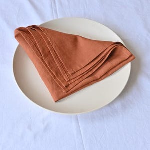 May include: A stack of folded, terracotta-coloured linen napkins rests on a cream-coloured plate. The plate is centered on a white, textured tablecloth. The napkins are neatly arranged, showcasing their soft texture and warm hue.