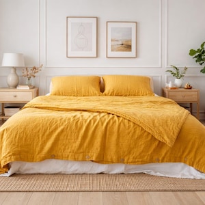 May include: A bedroom scene featuring a bed with a mustard-yellow linen duvet cover and matching pillows. The bed sits on a woven rug, with two wooden nightstands on either side. Artwork hangs above the bed, and a potted plant adds a touch of greenery.
