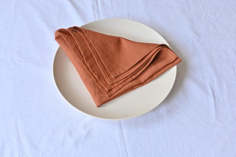 May include: A stack of folded, rust-colored linen napkins rests on a cream-colored plate. The plate is centered on a white, textured tablecloth. The napkins are neatly arranged, showcasing their soft texture and warm hue.