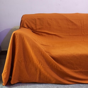 Op de afbeelding: Een oranje bankhoes over een bank gedrapeerd. De hoes is gemaakt van een zachte, getextureerde stof en heeft een warme, uitnodigende kleur. De bank staat tegen een witte muur en de vloer is een gespikkelde grijze tegel.