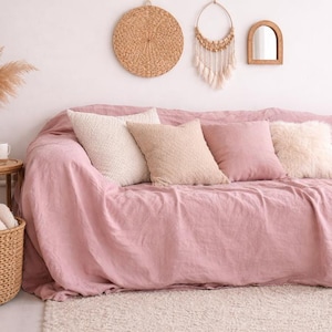 Puede incluir: Un salón acogedor con un sofá rosa y cojines decorativos. Una cesta de mimbre y una pequeña mesa auxiliar con un jarrón y tazas complementan el ambiente. Decoraciones de pared añaden encanto a la habitación.