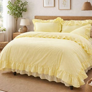 Può includere: Un letto con un copripiumino giallo con volant e federe abbinate. Il letto ha una testiera intrecciata ed è posto contro un muro dai toni neutri. Due stampe botaniche incorniciate sono appese sopra il letto.
