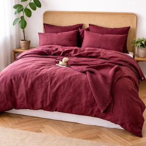 May include: A bed with a burgundy linen duvet cover, matching pillowcases, and a knitted throw. The bed has a woven headboard and sits on a wooden floor with a beige rug. Potted plants are on either side of the bed.
