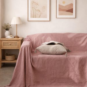 Könnte beinhalten: Ein Wohnzimmer mit einem staubrosafarbenen Sofabezug aus Leinen, einem beigen Kissen mit Quasten und zwei gerahmten Landschaftsdrucken. Ein hölzerner Beistelltisch mit einer Lampe und einem Weidenkorb ist ebenfalls zu sehen.