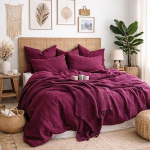 May include: A bedroom with a bed dressed in a burgundy duvet and pillows. A woven headboard and pendant light are visible. The room is decorated with a jute rug, macrame wall art, and framed pictures, creating a cozy atmosphere.