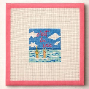 May include: Needlepoint art piece with a pink border and a central scene depicting a beach with the phrase "c'est la vie" in red script. The scene includes blue sky, white clouds, and figures in the water.