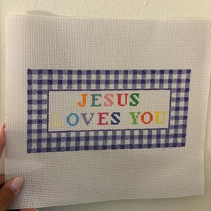 May include: Needlepoint canvas with the text "JESUS LOVES YOU" in colorful letters. The text is inside a blue and white gingham border. The canvas is white and held by a person.