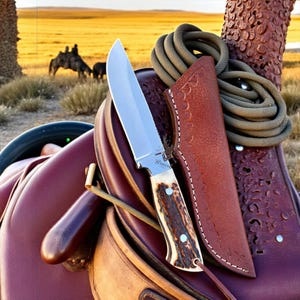 May include: A hunting knife with a stainless steel blade and a textured antler handle, resting on a burgundy leather saddle. The knife is in a brown leather sheath, with a coiled rope and a blurred background of a field and horses.