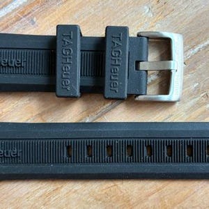 Puede incluir: Correa de reloj de goma negra con el nombre de la marca "TAGHeuer" en relieve. La correa tiene una hebilla plateada y está diseñada para un reloj. La correa está sobre una superficie de madera.
