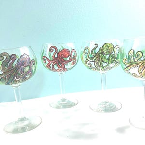 Puede incluir: Cuatro copas de cristal transparente con tallo, cada una con un pulpo pintado a mano en un color diferente: morado, rojo, verde y dorado. Cada pulpo se muestra con tentáculos detallados y rodeado de detalles de algas verdes.