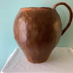 Op de afbeelding: Een grote, bruine, handgemaakte keramische kruik met een afgeronde body en een gebogen handvat. De kruik heeft een brede opening en een getextureerd oppervlak, wat wijst op een rustieke of antieke stijl. De rand is een lichtere bruintint.