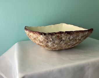 May include: A handcrafted, textured bowl with a rough, earthy exterior in shades of brown, beige, and white. The rim is a darker brown, and the interior is a pale yellow. The bowl is placed on a white surface.