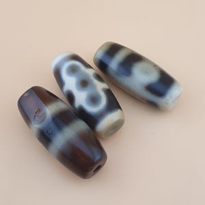 May include: Three elongated dzi beads with a mix of brown, white, and grey patterns. The beads have a smooth, polished appearance and are arranged on a light beige surface. The dzi beads are a type of Tibetan bead.
