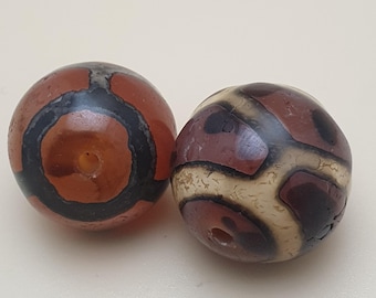 May include: Two round, patterned beads. One bead is primarily reddish-brown with a black ring and a central hole. The other bead has a pattern of dark brown, beige, and reddish-brown shapes. Both beads have a hole.