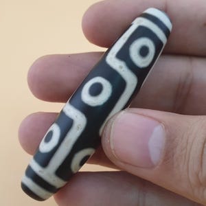 May include: A black and white dzi bead with a cylindrical shape. The bead features a pattern of white circles and lines against a black background. The bead is held in a hand.