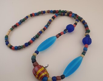May include: A vibrant beaded necklace with a mix of bead shapes and colours. The necklace features small, round beads in various colours, with larger blue oval and round beads. A patterned clasp fastens the necklace.