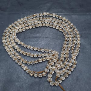 May include: A necklace composed of multiple strands of clear, round beads with a light brown hue. The beads are strung together with a brown cord, creating a layered effect. The necklace is displayed against a dark background.